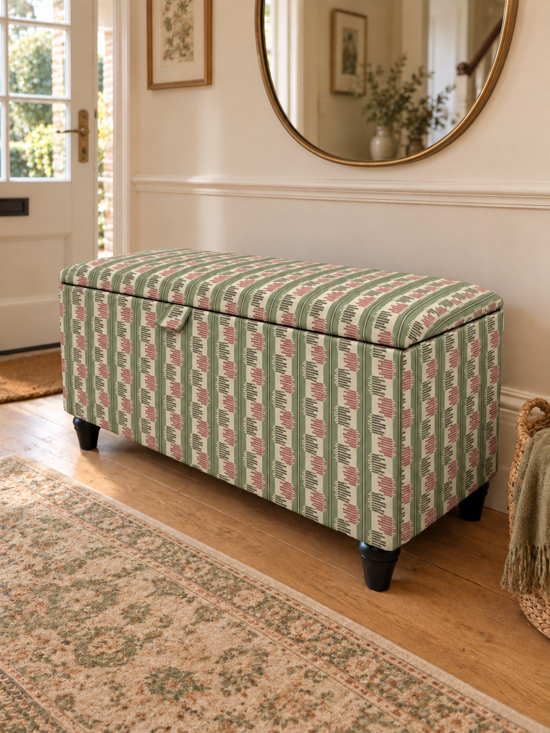 An upholstered storage bench with a green, pink, and white striped patterned fabric, set in a bright entryway on a wooden floor next to a patterned rug.