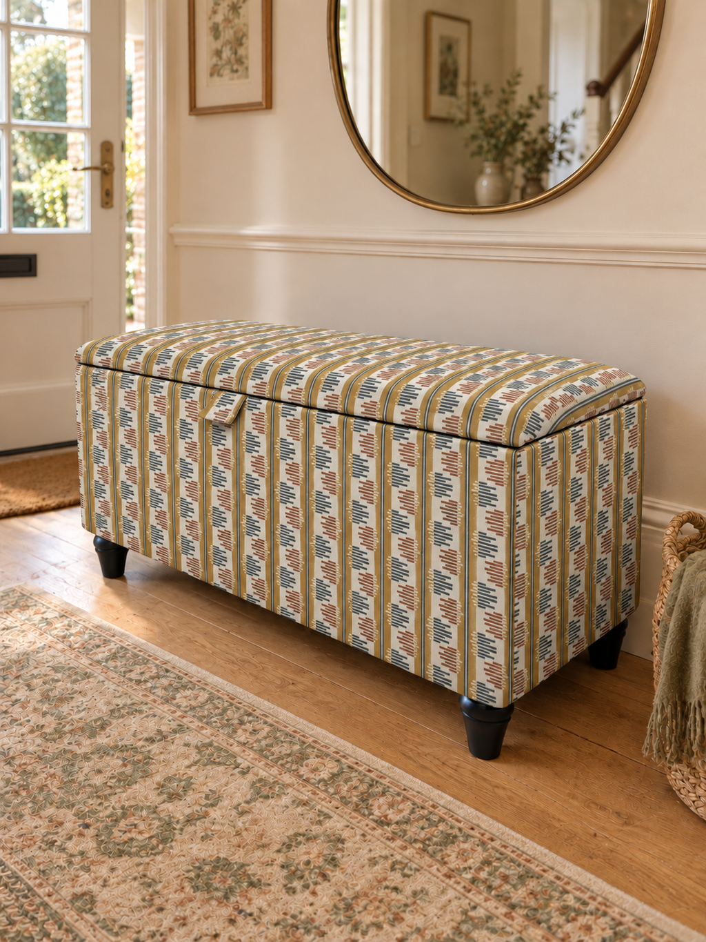 An upholstered storage bench with a striped patterned fabric in beige, green, and blue, set in a bright entryway with a wooden floor and a large round mirror on the wall.