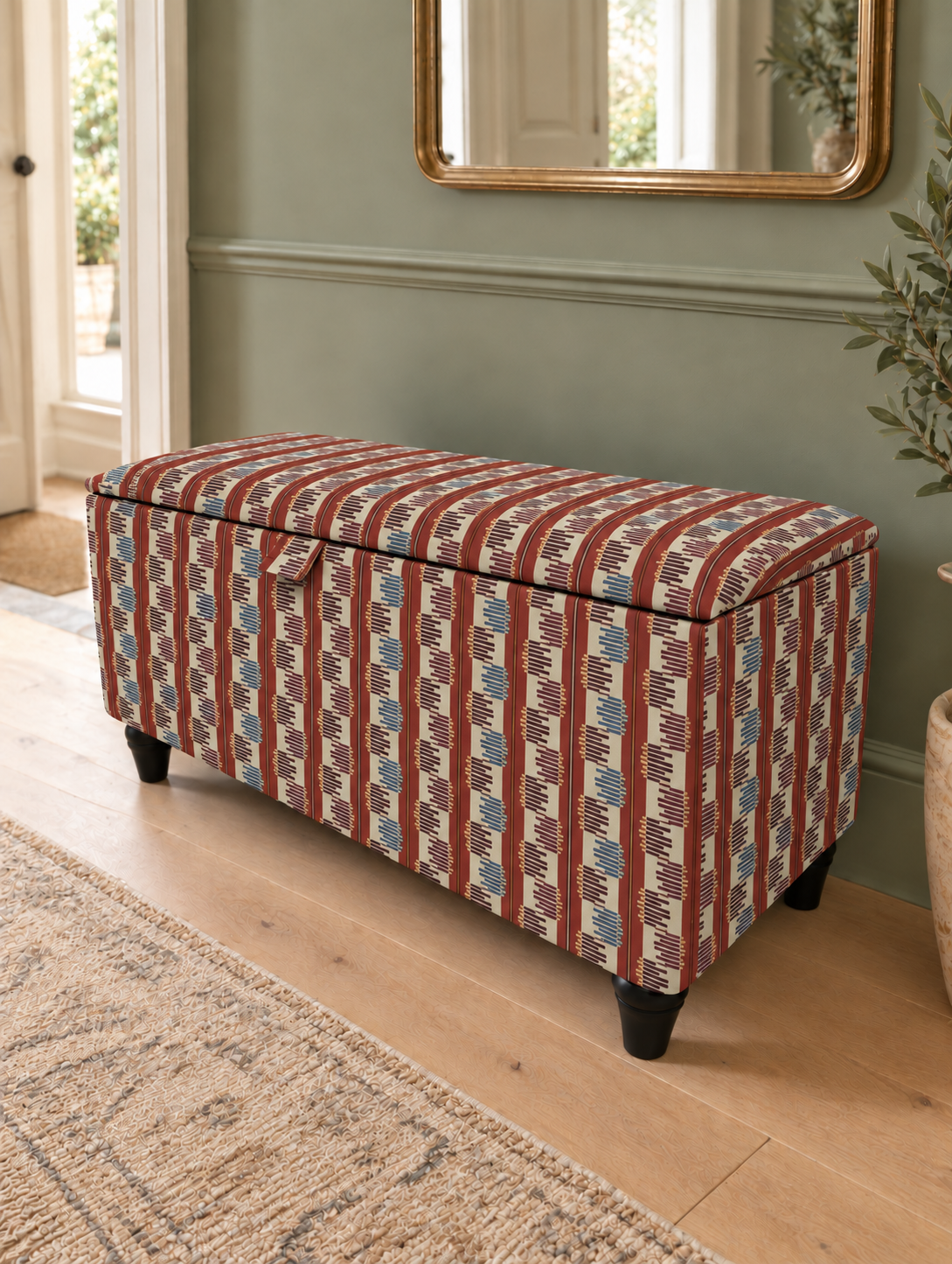 An upholstered storage bench with a geometric patterned fabric in rust red, cream, and blue, set in a room with light wood floors and a sage green wall.