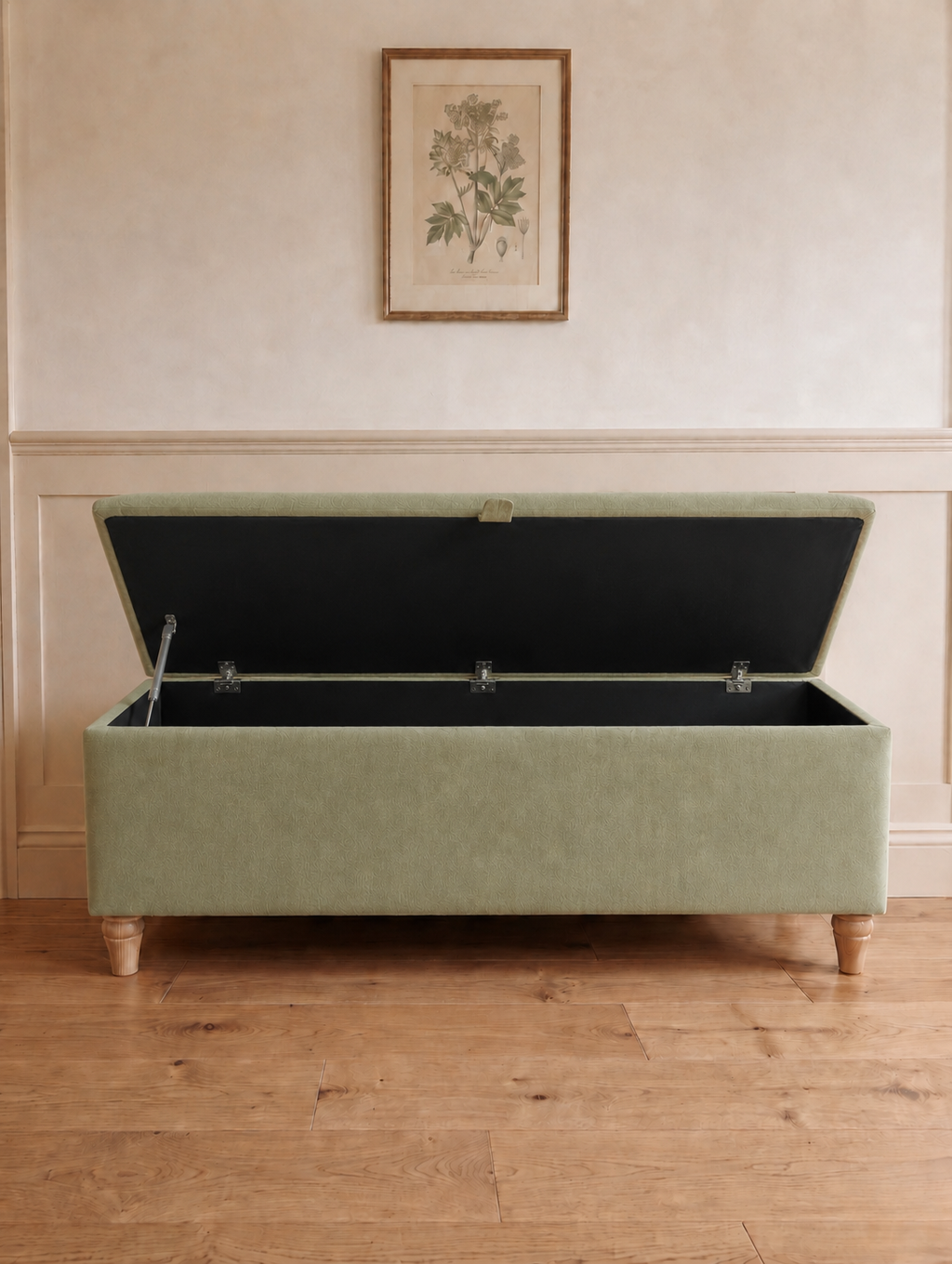 A rectangular upholstered storage bench in a muted sage green fabric open with black lining with turned wooden legs, set against a cream paneled wall with a framed botanical print above.