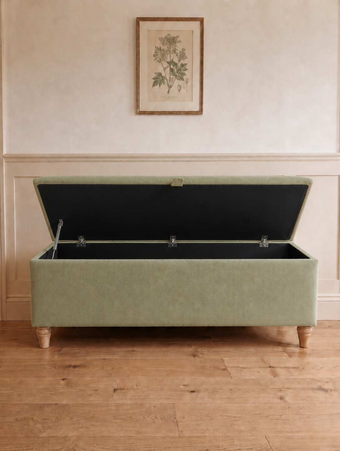 A rectangular upholstered storage bench in a muted sage green fabric open with black lining with turned wooden legs, set against a cream paneled wall with a framed botanical print above.