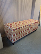 Upholstered storage bench in a Red and gold Paxton stripe and checked fabric by James Hare. Sat on a workshop floor with pink panelled walls behind. 