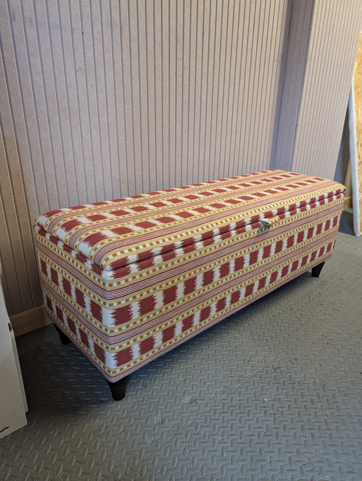 Upholstered storage bench in a Red and gold Paxton stripe and checked fabric by James Hare. Sat on a workshop floor with pink panelled walls behind. 