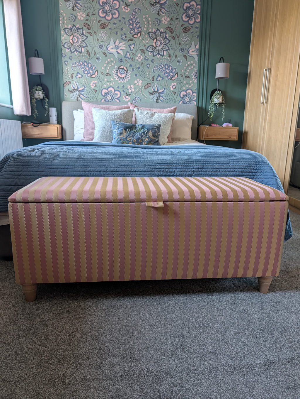 A striped fabric storage bench with a lid sits at the foot of a bed in a styled bedroom. The bench features vertical stripes in dusty pink and beige.