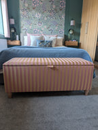 A striped fabric storage bench with a lid sits at the foot of a bed in a styled bedroom. The bench features vertical stripes in dusty pink and beige.
