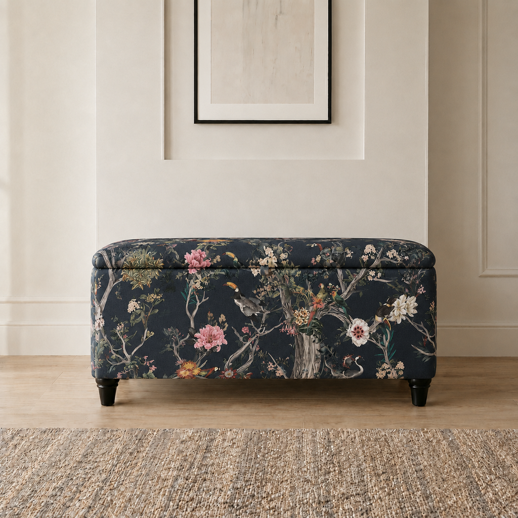 A rectangular upholstered storage bench with a dark navy floral and bird pattern, standing on a light wood floor in front of a cream paneled wall.