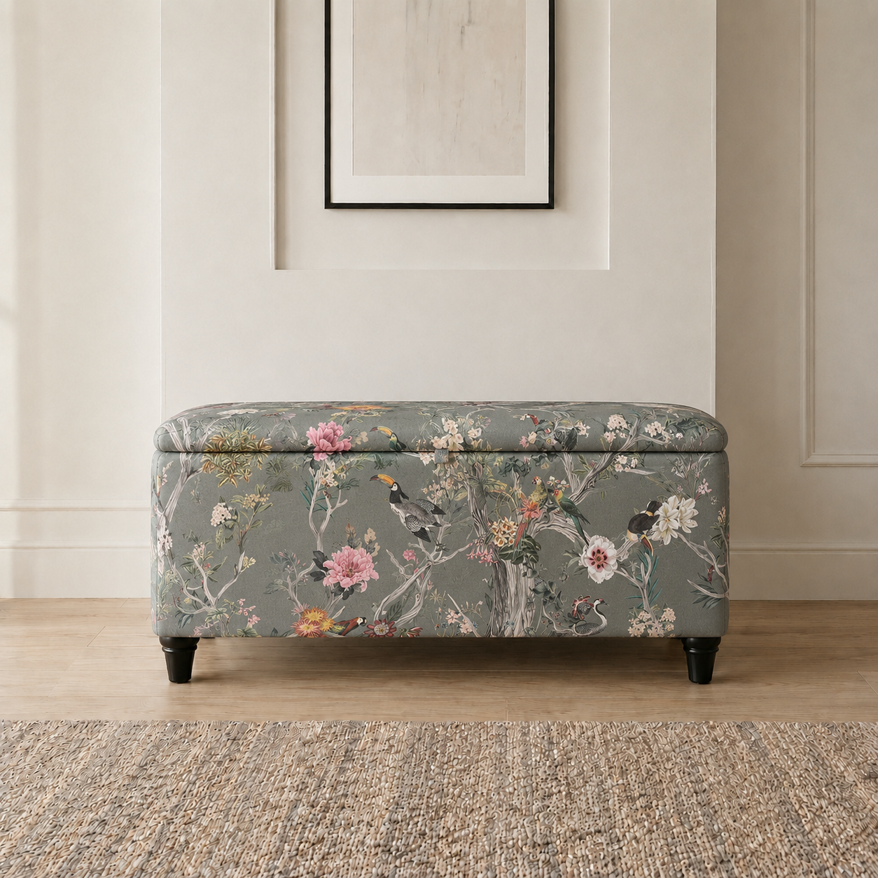A rectangular upholstered storage bench with a floral and bird pattern on a muted sage green background, positioned on a light wood floor in front of a cream paneled wall.