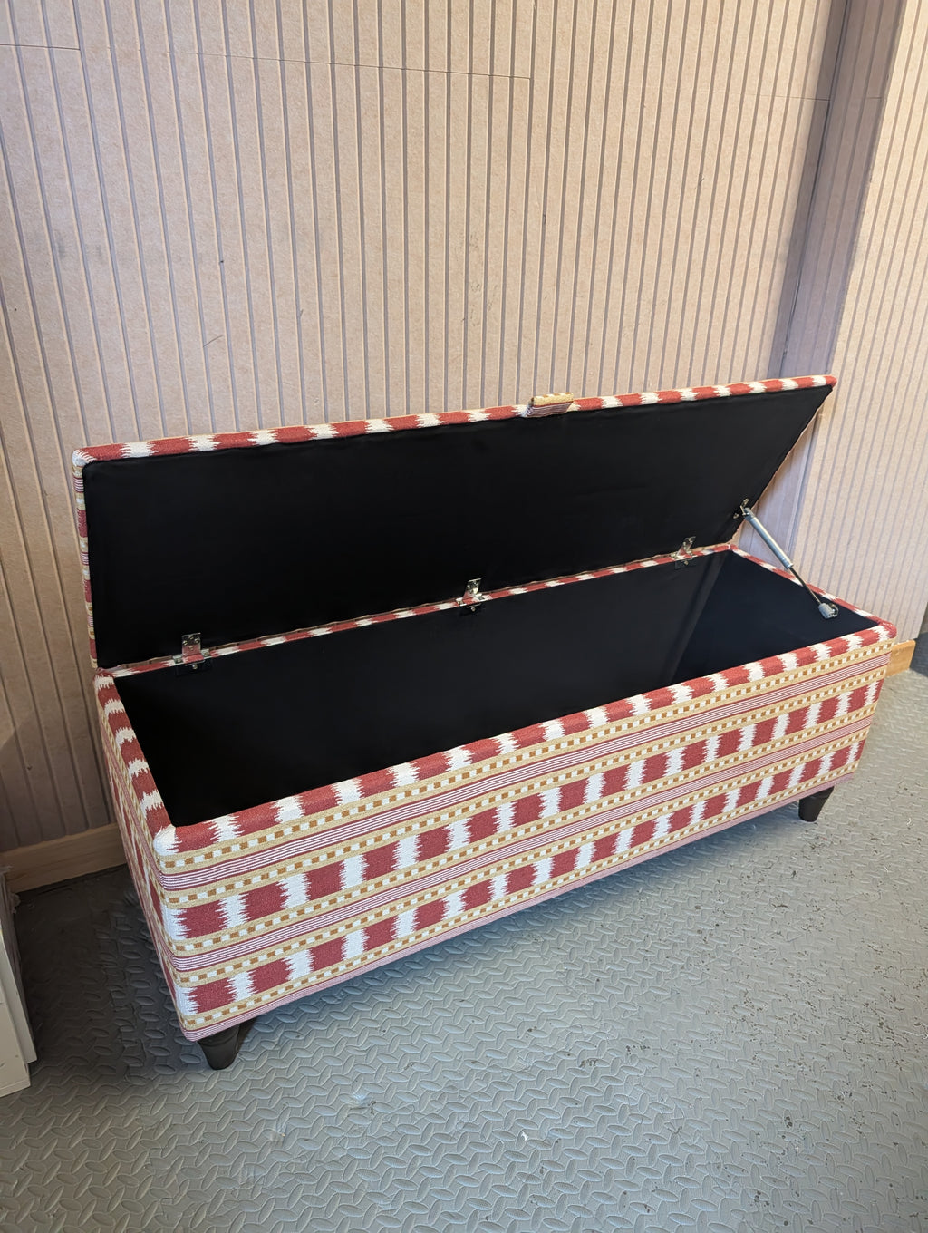A rectangular storage bench with a patterned fabric upholstery in red, white, and gold, shown with its lid open to reveal a black interior. The bench is positioned on a gray textured floor against a beige paneled wall.