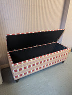 A rectangular storage bench with a patterned fabric upholstery in red, white, and gold, shown with its lid open to reveal a black interior. The bench is positioned on a gray textured floor against a beige paneled wall.