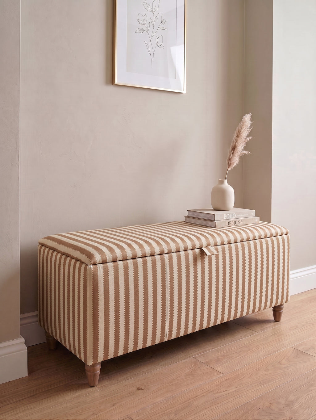 A rectangular storage bench with a cream and brown regency striped fabric upholstery, styled in a home entryway with a wooden floor, a vase with dried grass, and a framed print on the wall.
