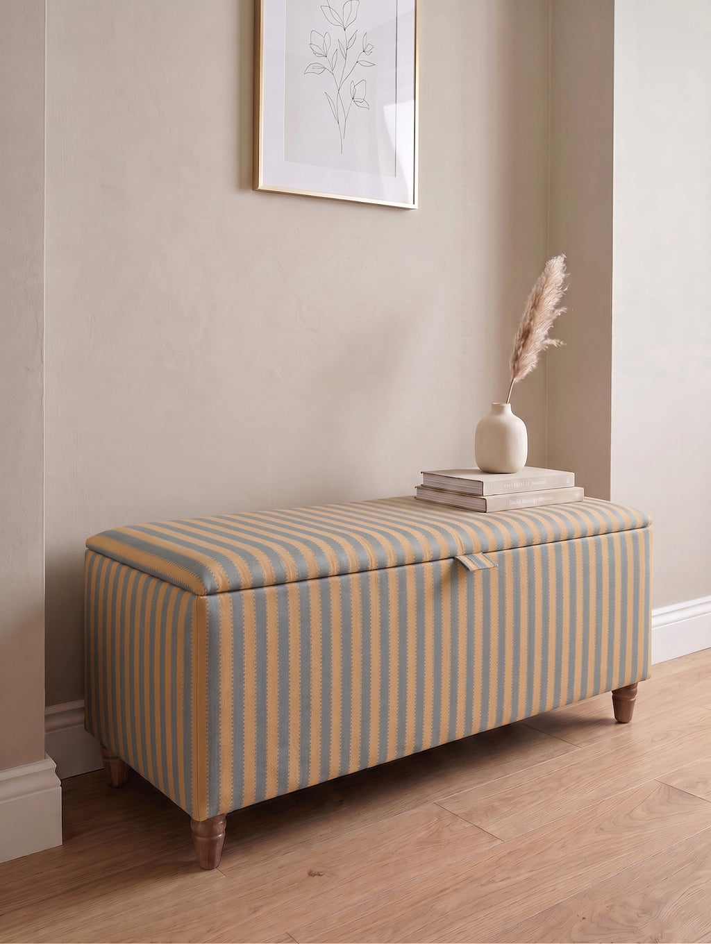 A striped fabric storage bench with a hinged lid, styled with a small stack of books and a vase of dried grass on top, set against a beige wall and light wood floor.