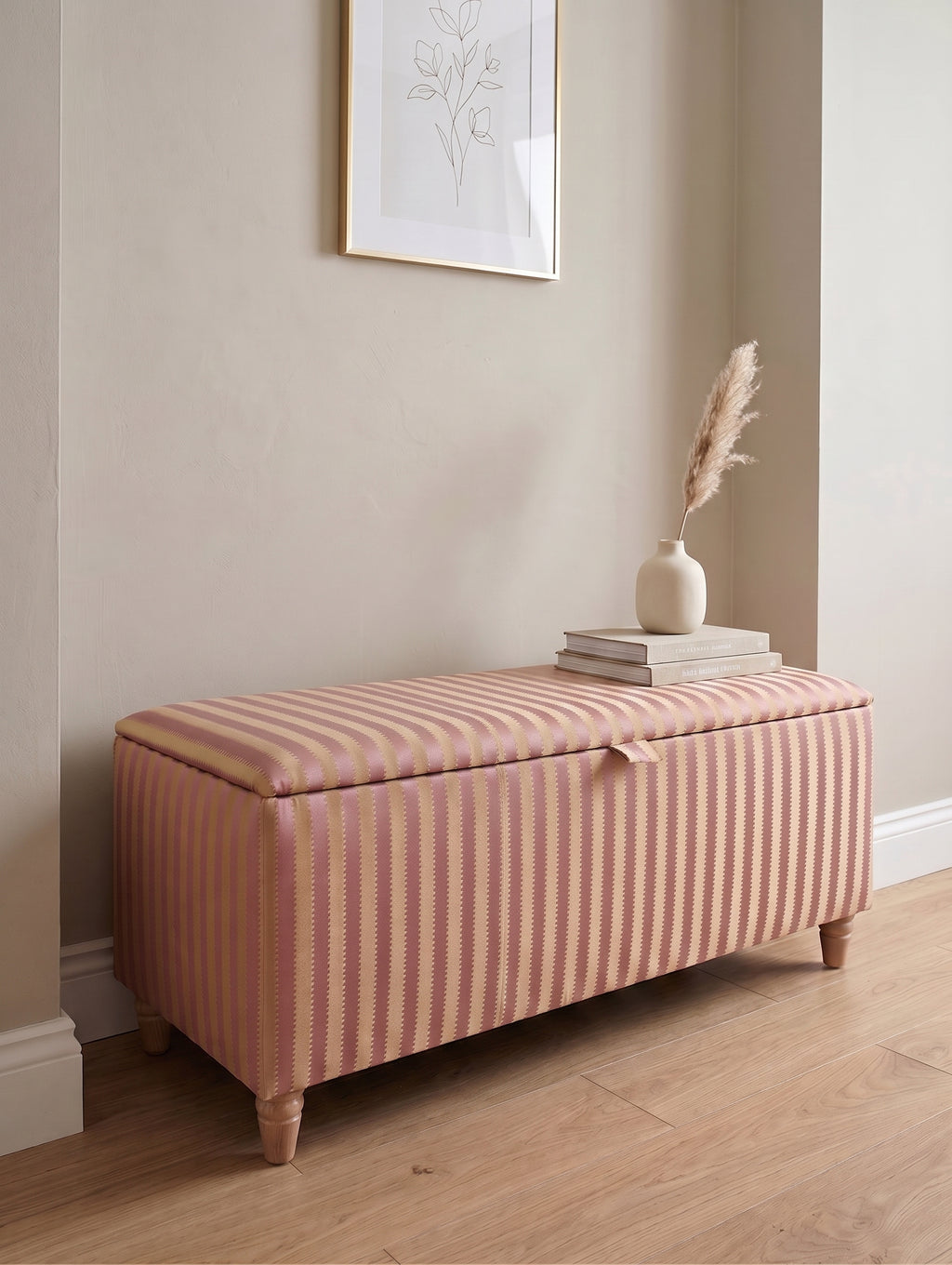 A rectangular storage bench with a hinged lid upholstered in pink and yellow Sanderson regency vertical stripes, styled in a room corner with a wooden floor, a vase with dried grass, and a framed print on the wall.