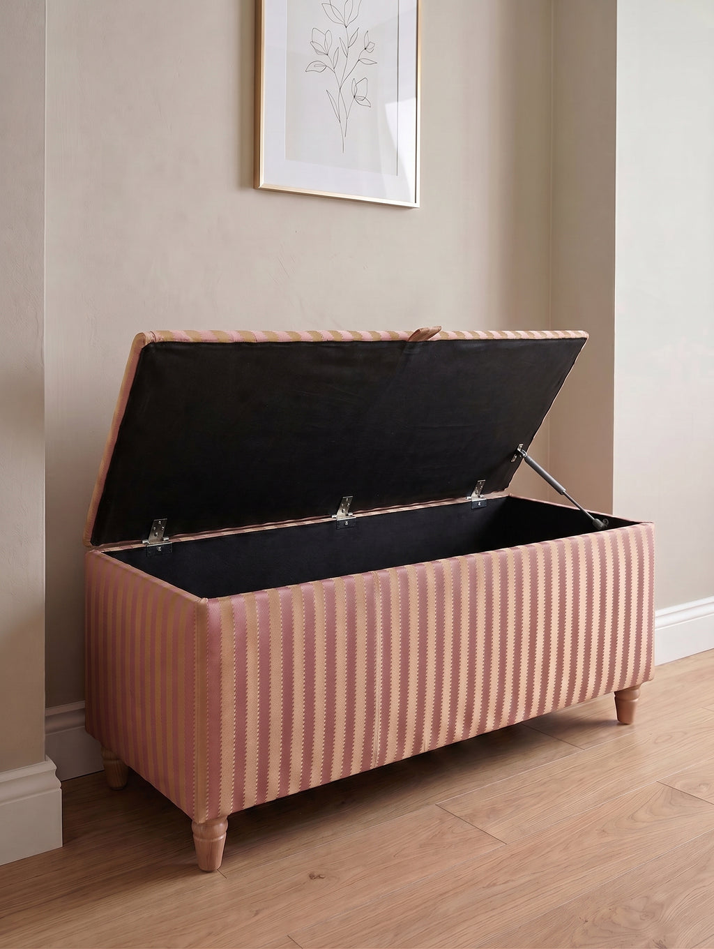 A pink and yellow Sanderson regency vertically striped fabric storage bench with wooden legs, shown with its lid open to reveal a black cushioned interior, set in a room with light wood floors and beige walls.