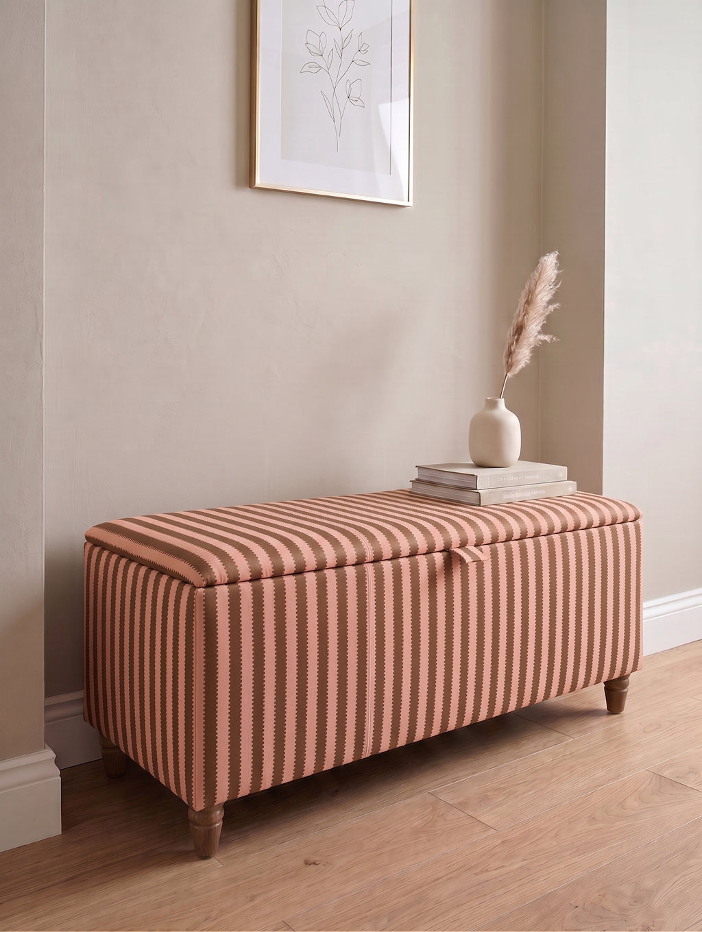 A rectangular storage bench with a Sanderson regency striped fabric upholstery in pink and brown, styled in a home entryway with a wooden floor, books, a small vase, and a framed print on the wall.