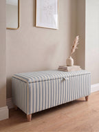 A striped fabric storage bench with a hinged lid, styled in a home entryway with a stack of books and a vase on top.