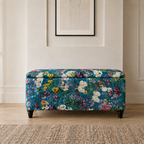 A rectangular upholstered storage bench with a vibrant multicolor floral pattern on a teal background, featuring dark wooden legs, set in a styled room with a light wood floor and a neutral wall.