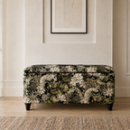 A rectangular upholstered bench with a dark floral and botanical pattern in black, cream, and olive green, standing on short dark legs in a styled interior setting.