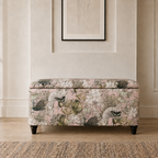 A rectangular upholstered storage bench with a floral and bird pattern in soft pink, beige, and green, set against a neutral cream wall with a framed print above.