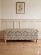 Decorative floral-patterned ottoman on a wooden floor with panelled walls and a framed picture.