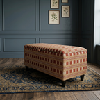 A maroon, red and orange modern box stripped storage ottoman on a vintage style rug in a room with dark panelled walls.