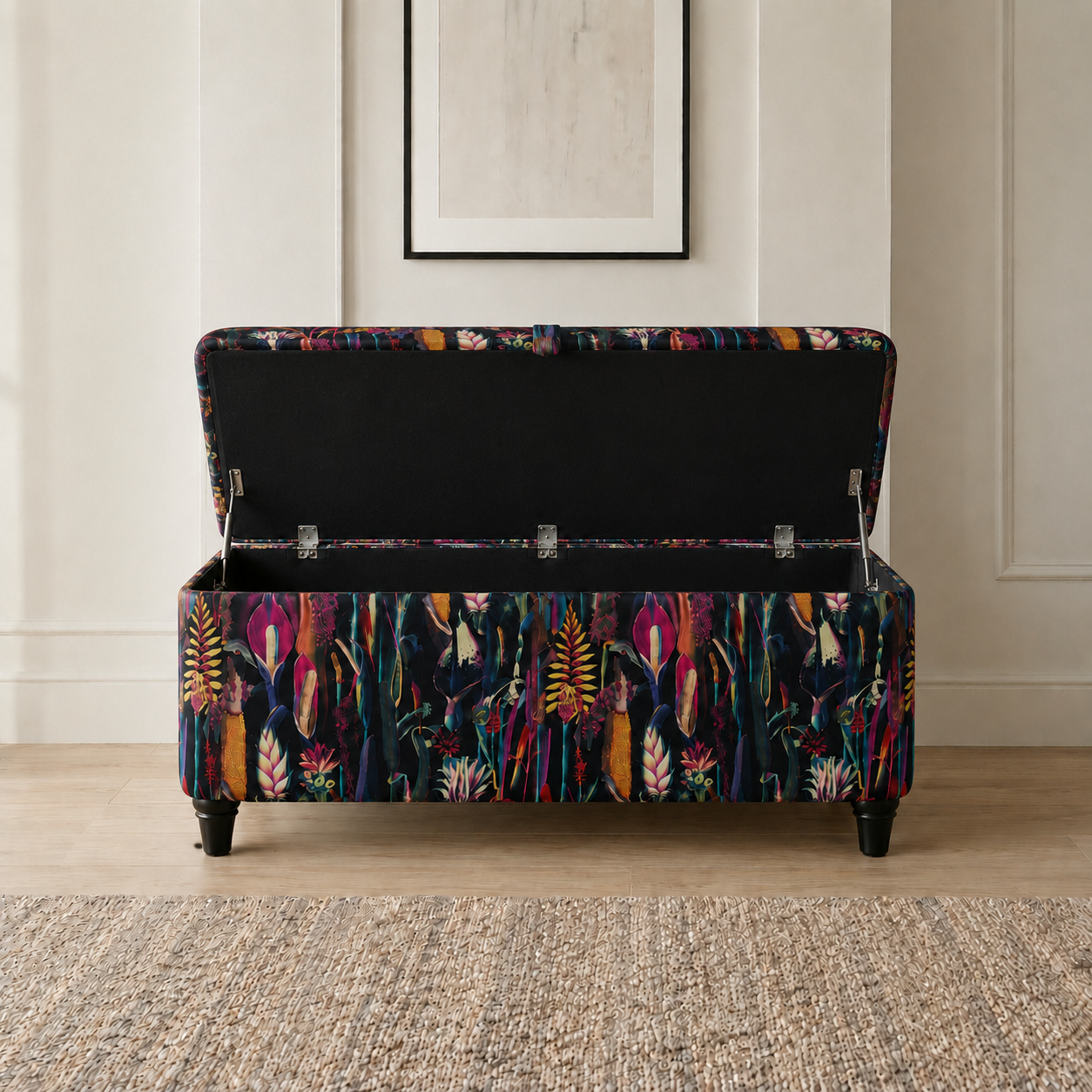 A storage bench with a colorful tropical floral patterned upholstery, shown open to reveal a black fabric-lined interior. The bench is positioned on a textured beige rug in a room with light wood floors and a neutral wall with a framed picture.