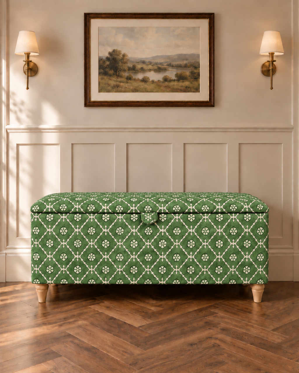 A green and white patterned fabric storage bench with wooden legs, set against a white paneled wall with a framed landscape painting and two wall sconces.