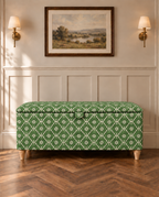A green and white patterned fabric storage bench with wooden legs, set against a white paneled wall with a framed landscape painting and two wall sconces.