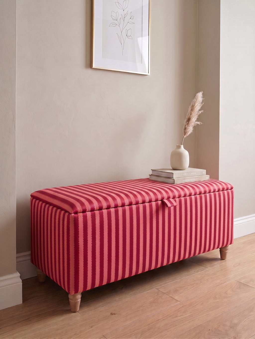 A rectangular storage bench with a red and pink Sanderson regency vertically striped fabric upholstery and light wood legs, styled in a home entryway setting with a vase and books on top.