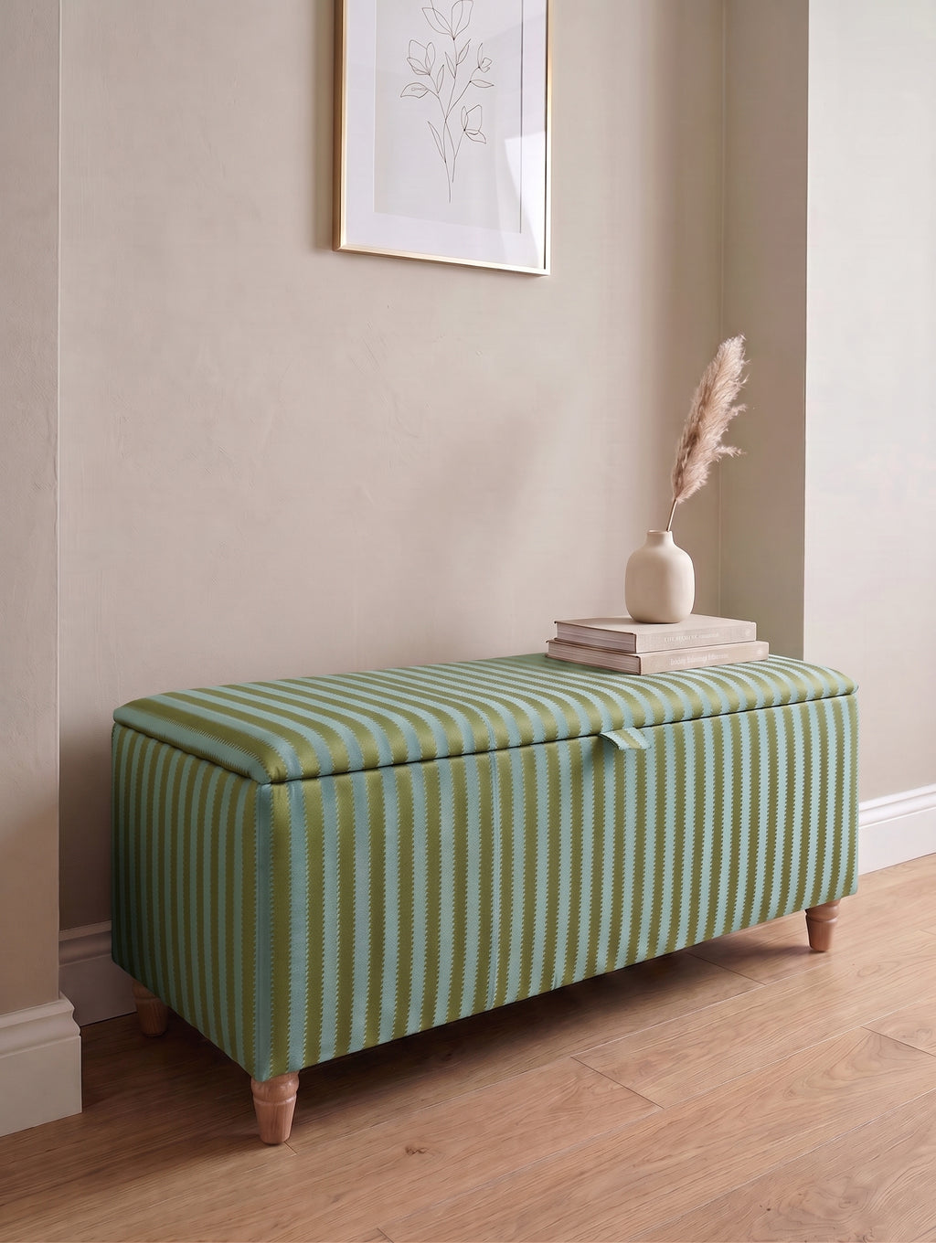 A storage bench with a green and light blue regency striped fabric upholstery, styled in a home entryway with a wooden floor, books, and a vase.