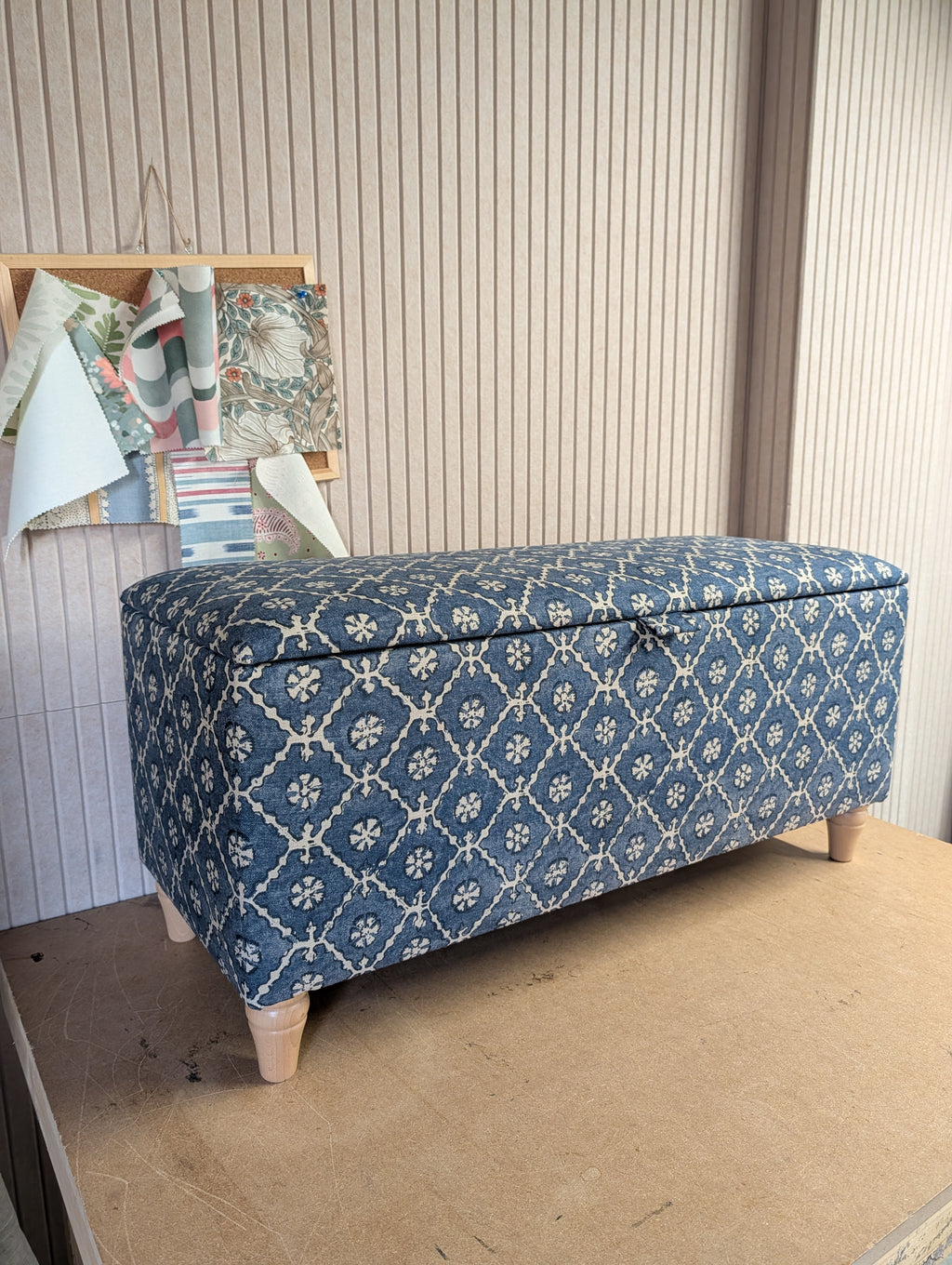 A rectangular storage bench with a hinged lid, upholstered in a blue and cream floral lattice pattern, standing on light-colored wooden legs against a beige paneled wall.