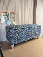 A rectangular storage bench with a hinged lid, upholstered in a blue and cream floral lattice pattern, standing on light-colored wooden legs against a beige paneled wall.