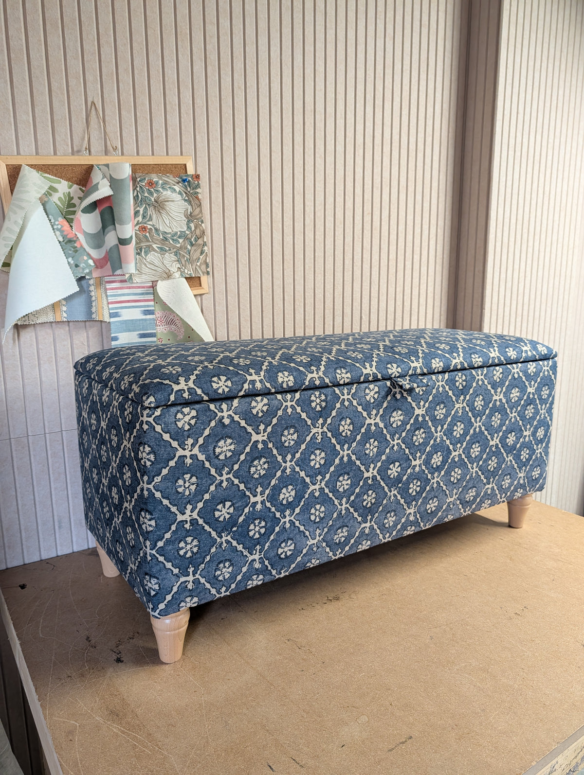 A rectangular storage bench with a hinged lid, upholstered in a blue and cream floral lattice pattern, standing on light-colored wooden legs against a beige paneled wall.