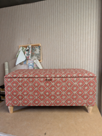 A storage bench with a patterned dusty rose and cream fabric, standing on light-colored wooden legs against a beige paneled wall.