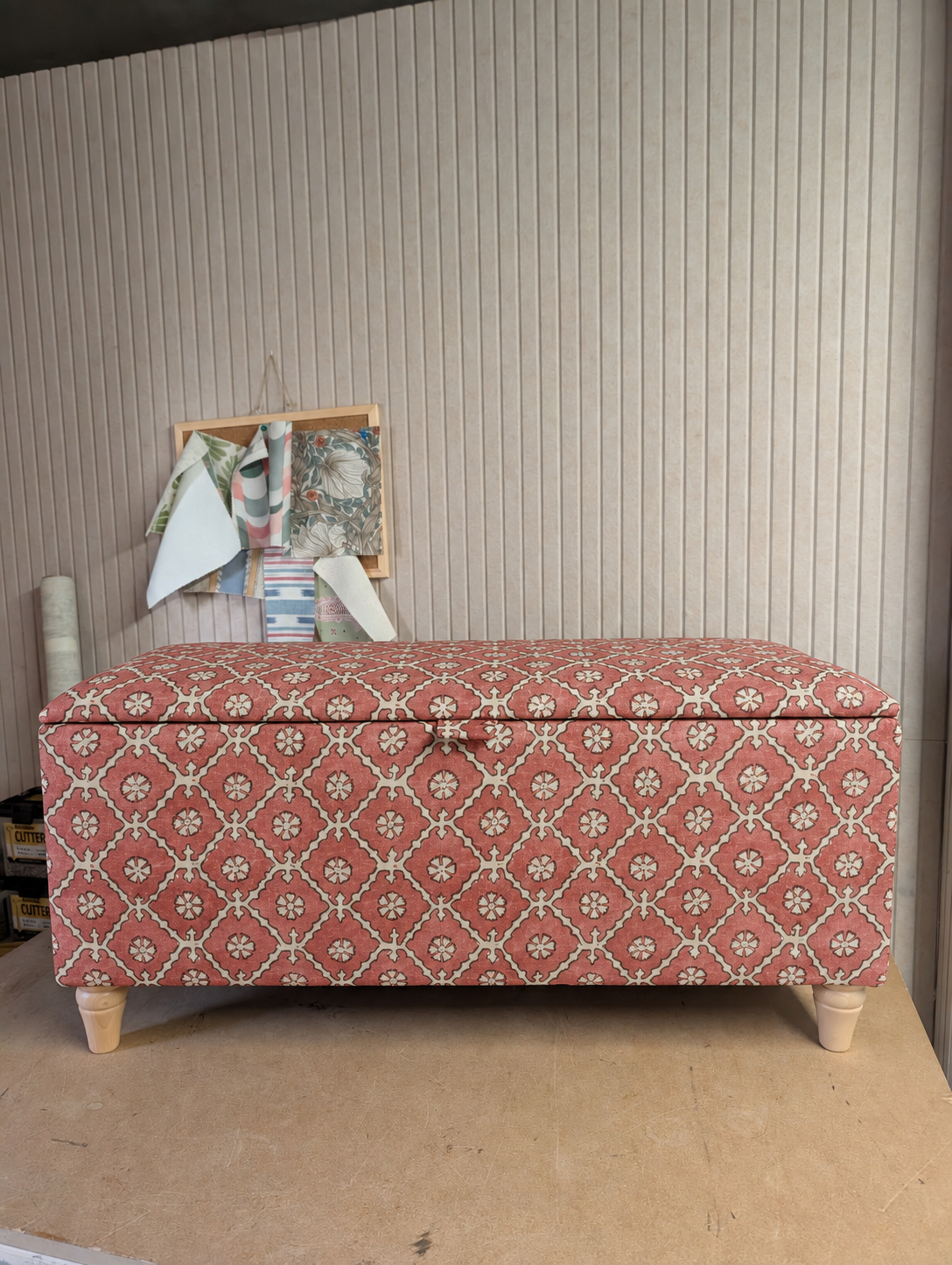 A storage bench with a patterned dusty rose and cream fabric, standing on light-colored wooden legs against a beige paneled wall.