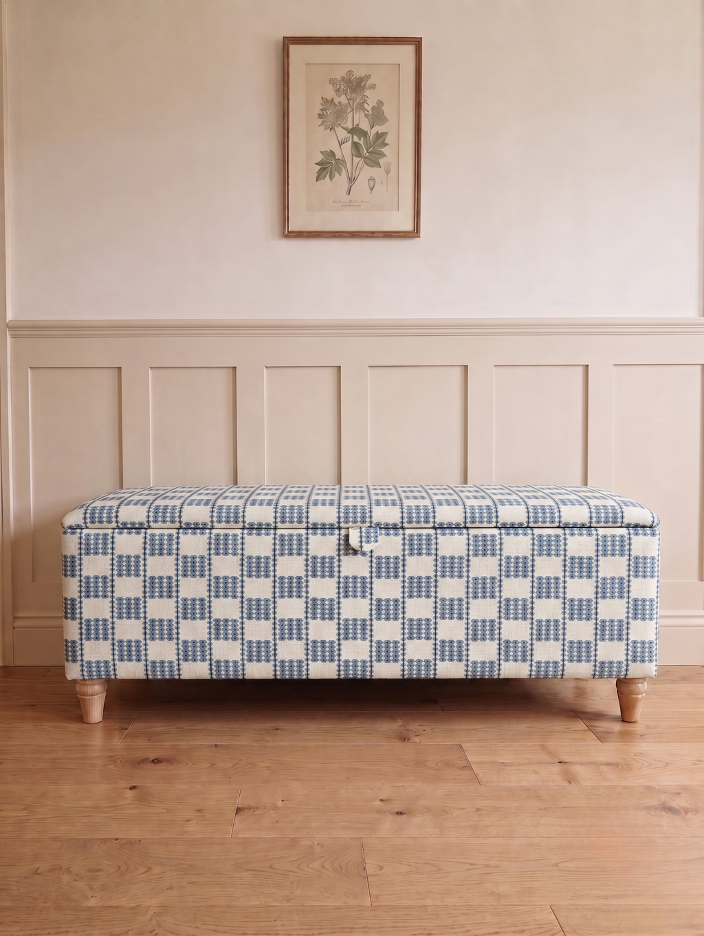 An upholstered storage bench with a blue and white checkered pattern, standing on wooden legs in a room with beige wainscoting and a light wood floor.