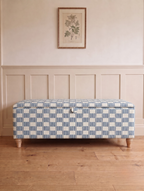An upholstered storage bench with a blue and white checkered pattern, standing on wooden legs in a room with beige wainscoting and a light wood floor.