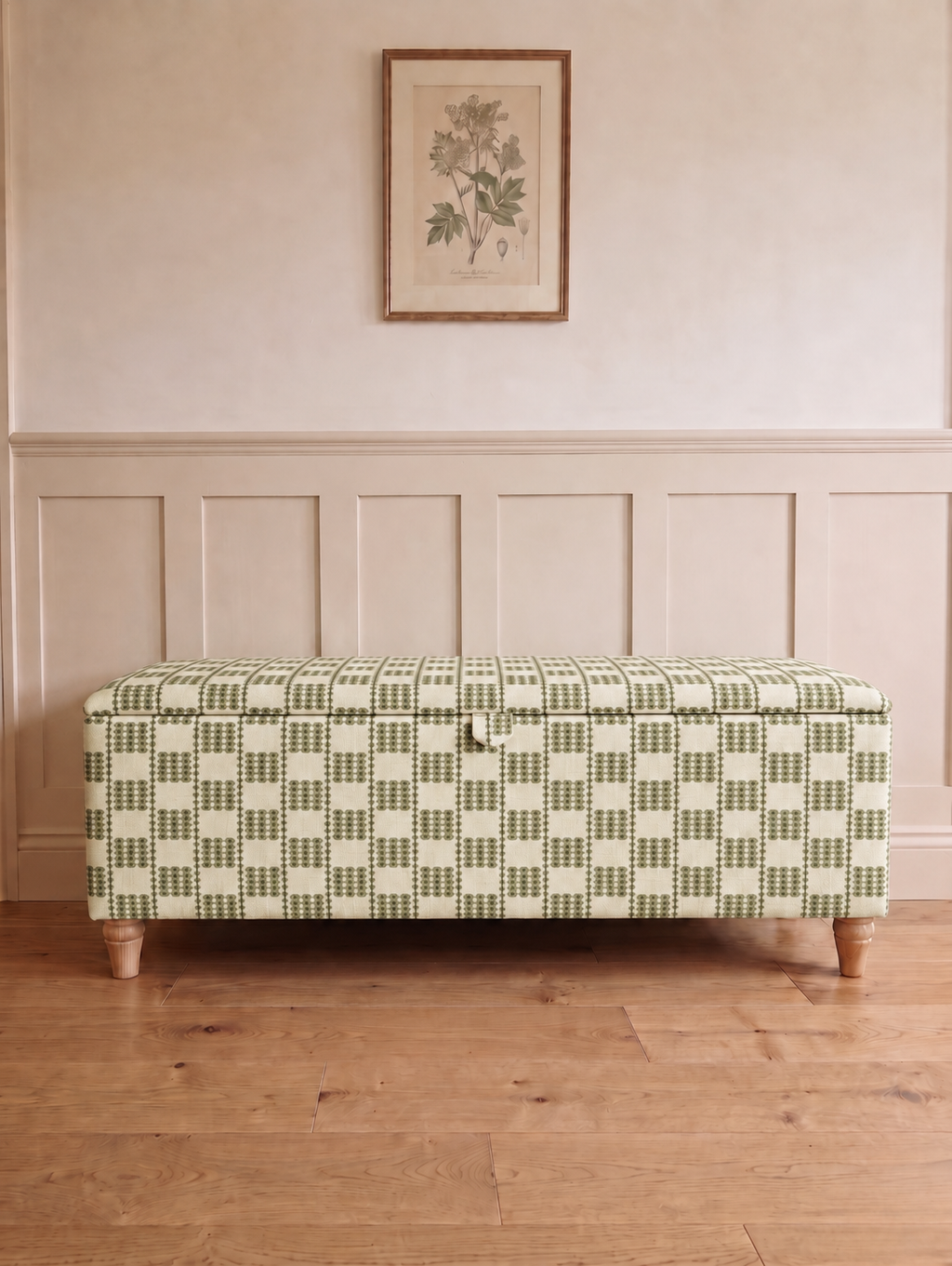 An upholstered storage bench with a green and cream geometric checkered pattern, positioned on a light wood floor against a beige paneled wall with a framed botanical print above.