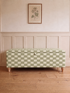 An upholstered storage bench with a green and cream geometric checkered pattern, positioned on a light wood floor against a beige paneled wall with a framed botanical print above.