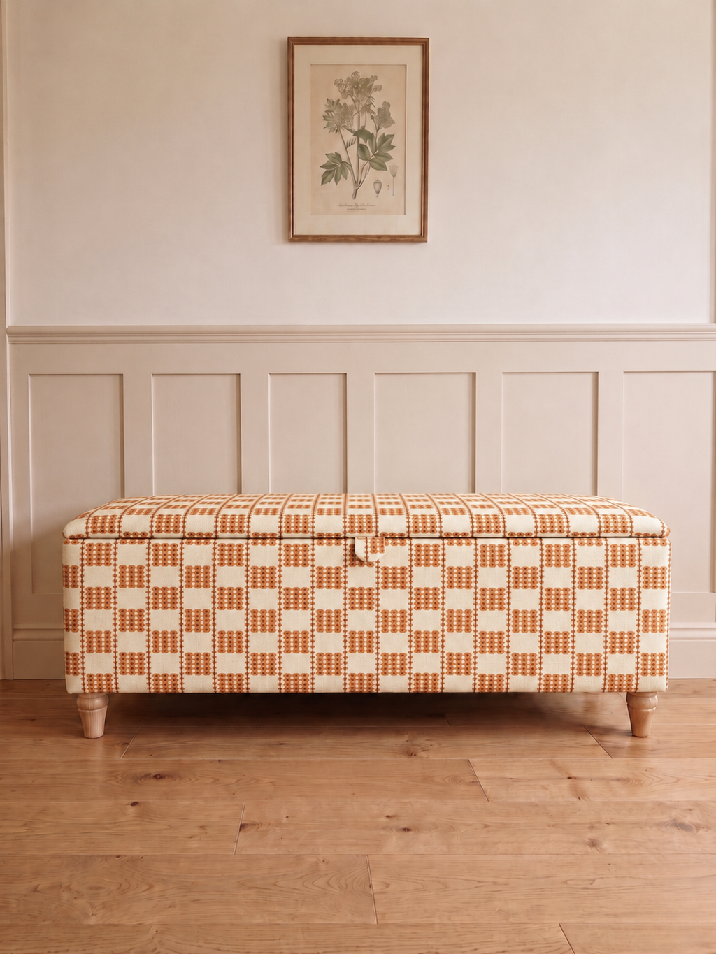 A storage bench upholstered in a warm orange and cream checkered pattern, positioned against a white panelled wall with a framed botanical print above and a light wood floor below.