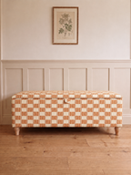 A storage bench upholstered in a warm orange and cream checkered pattern, positioned against a white panelled wall with a framed botanical print above and a light wood floor below.