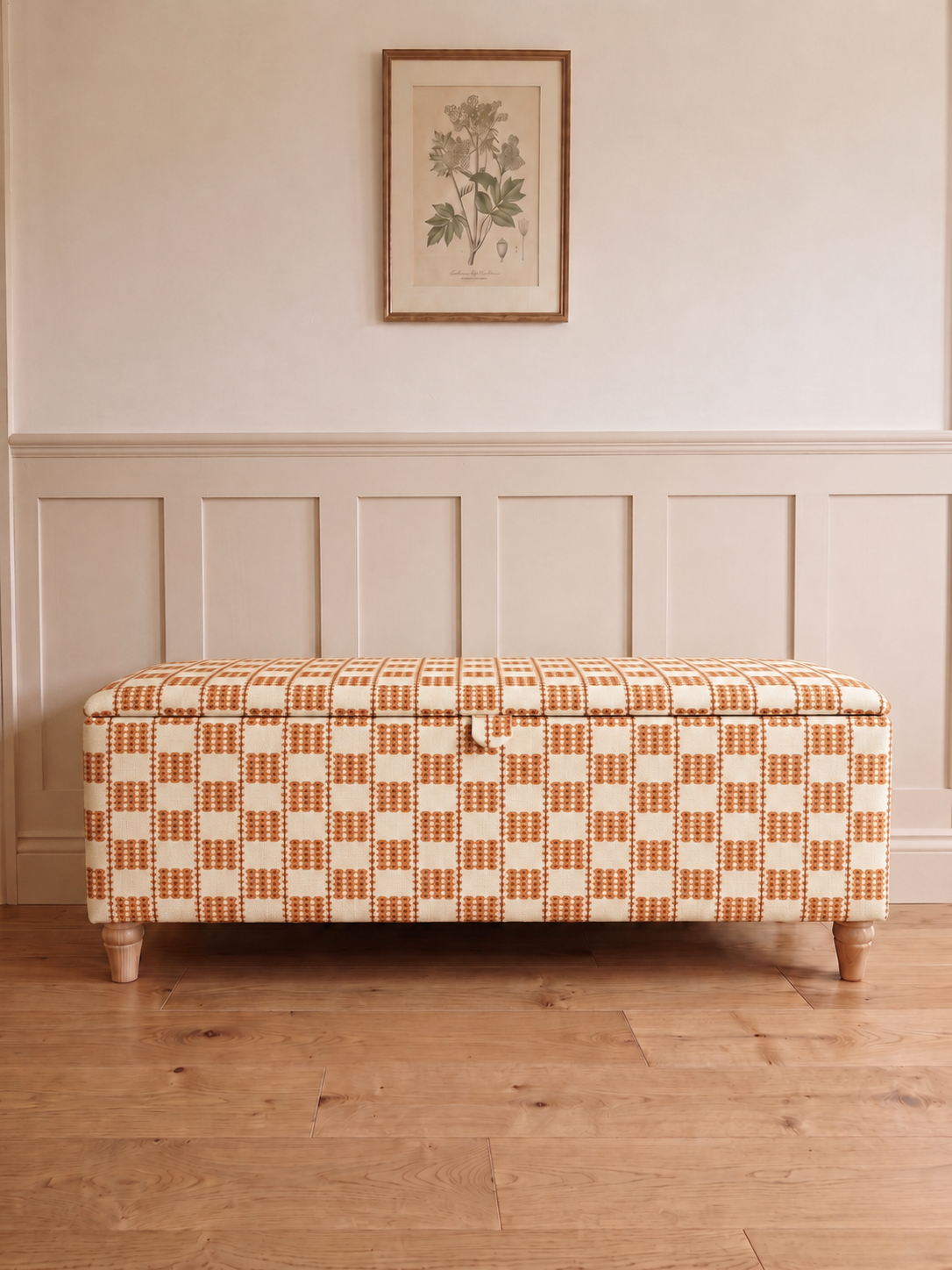 A storage bench upholstered in a warm orange and cream checkered pattern, positioned against a white panelled wall with a framed botanical print above and a light wood floor below.