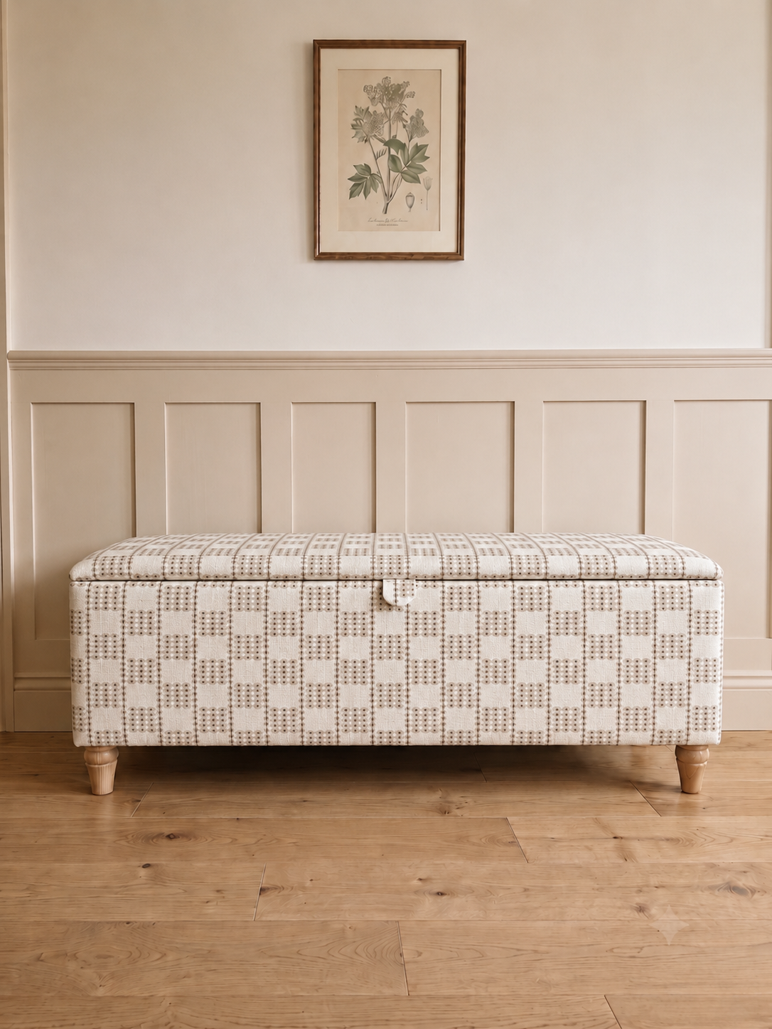 A storage bench with a cream and beige geometric basketweave pattern, featuring turned wood legs and a small front pull handle, set against a wainscoted wall with a framed botanical print.