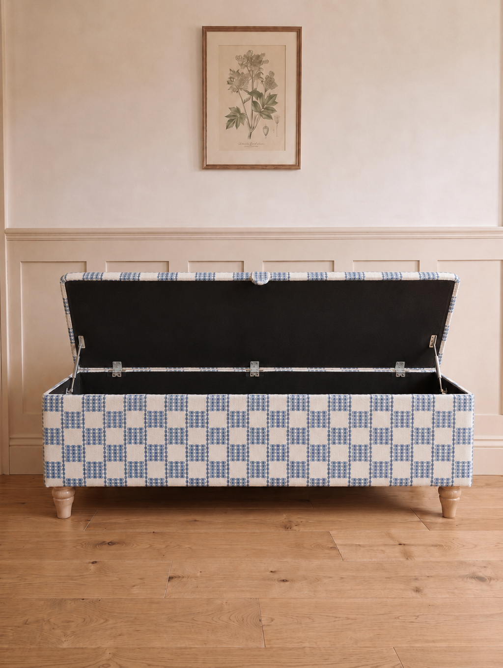 A storage bench with a blue and white checkered patterned fabric upholstery, shown with its lid open to reveal a black cushioned interior. The bench is positioned on a light wood floor against a cream-colored wall with wainscoting and a framed botanical print above.