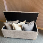 Grey bouclé fabric storage ottoman unit with open lid revealing cushions against a textured wall.