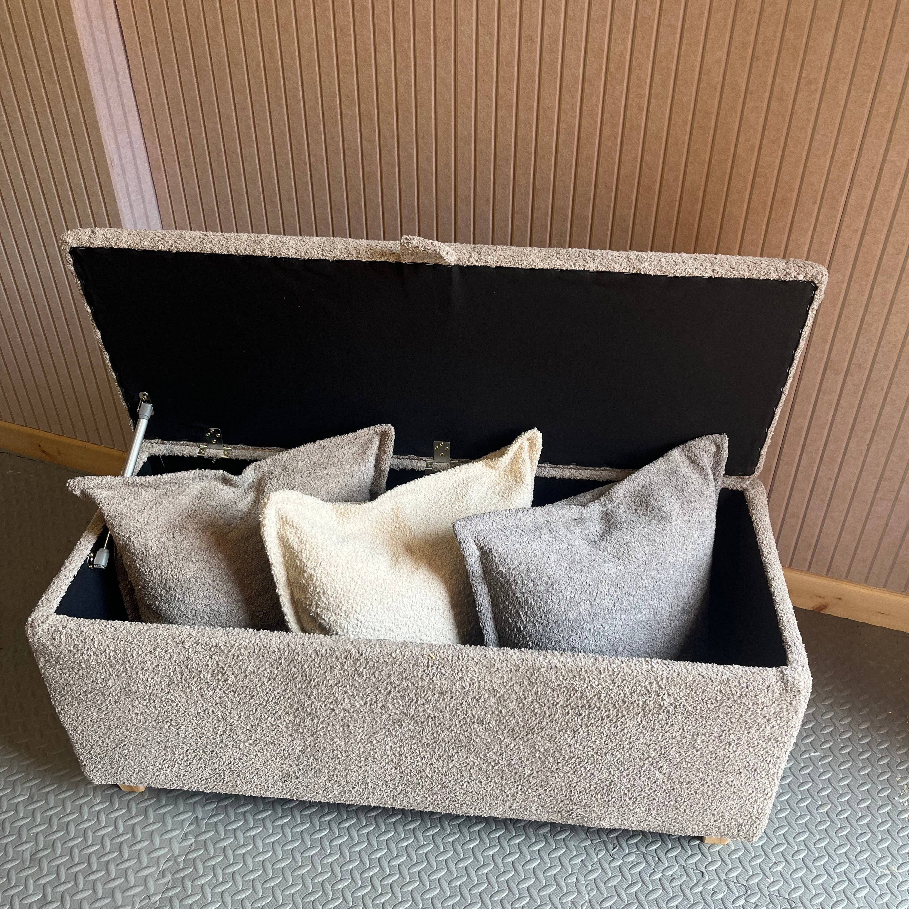 Grey bouclé fabric storage ottoman unit with open lid revealing cushions against a textured wall.