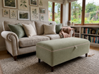 A styled living room scene featuring a light beige upholstered sofa with green and cream throw pillows, a matching sage green ottoman with pink piping in the foreground, and a bookshelf with framed botanical prints on the wall.
