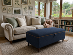 A navy blue upholstered ottoman with orange piping and dark wooden legs sits in front of a cream sofa in a bright, sunlit living room with bookshelves and botanical wall art.