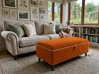 An orange upholstered rectangular ottoman with white piping and dark wooden legs sits in a bright living room in front of a cream sofa with green and beige pillows.
