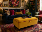 A styled living room featuring a deep navy blue velvet sofa with patterned and solid accent pillows, a mustard yellow upholstered ottoman coffee table with pink piping, and a large red and blue oriental rug.