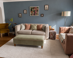 A styled living room featuring a light beige sofa with striped and textured throw pillows, a sage green velvet ottoman with pink piping, a mustard yellow armchair, and a pink armchair, set against a slate blue wall with framed botanical art.